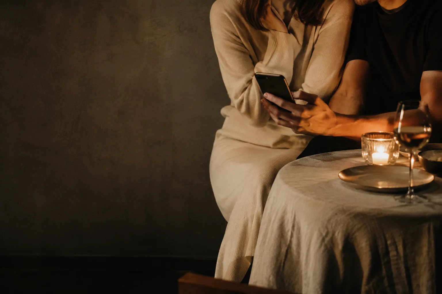 Two people seated close together in candlelight, with a phone and a blank card on the table.